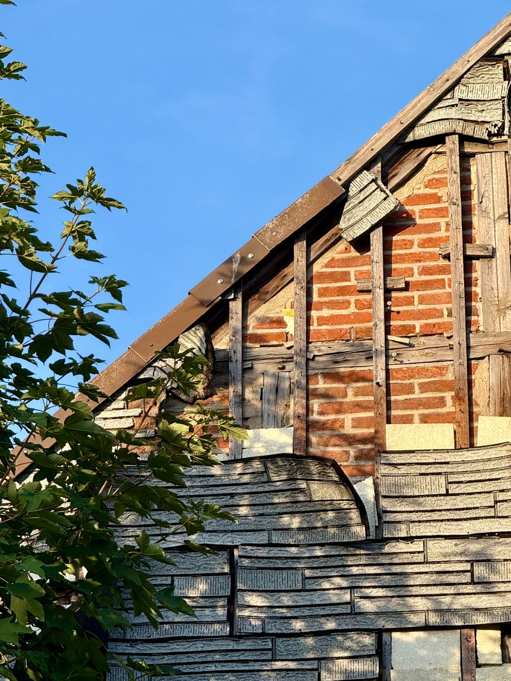 Pignon d’une vieille maison au revêtement usé et arraché, laissant voir la brique et la charpente.