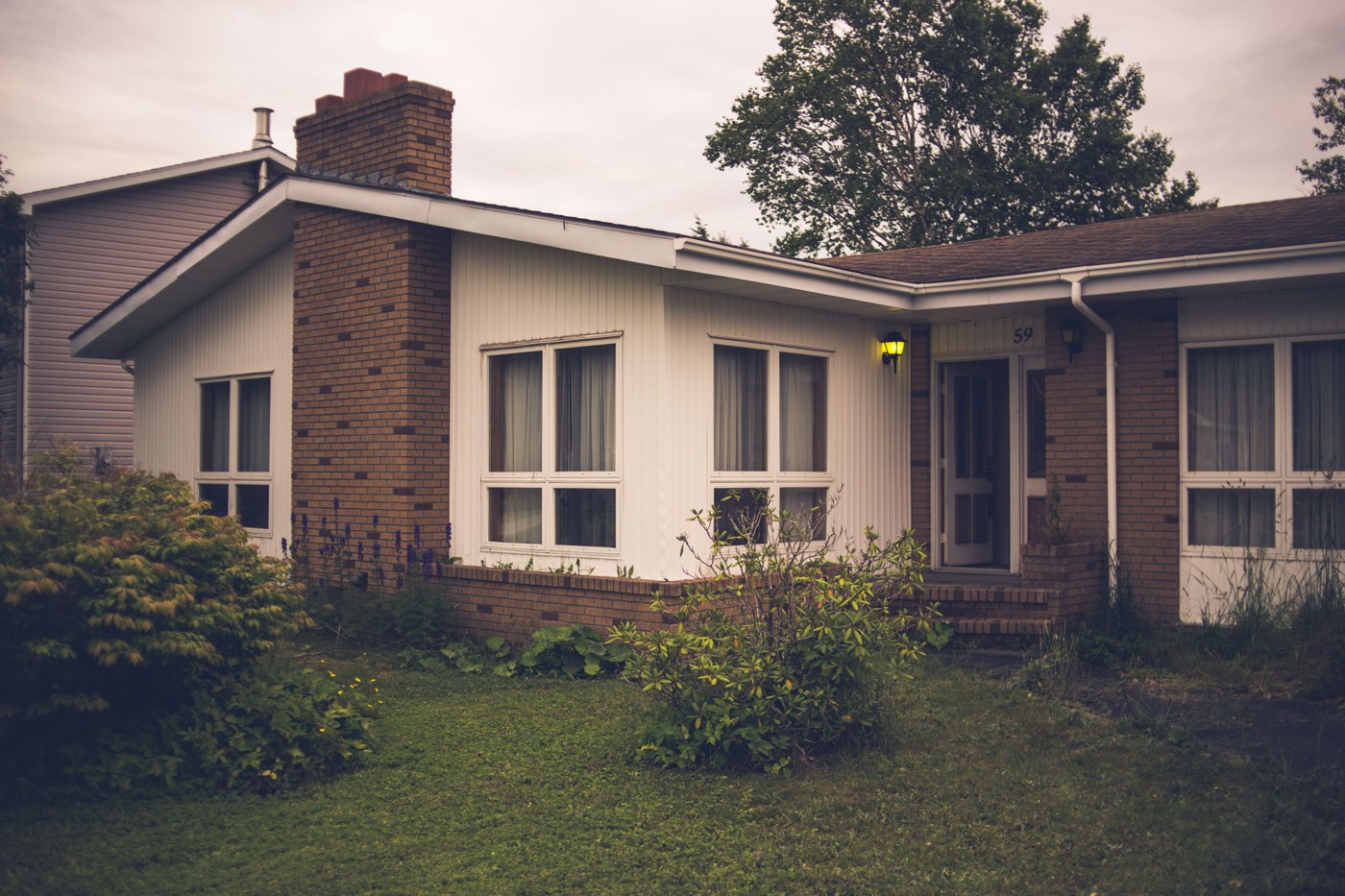 Bungalow des années 80 avec cheminée de brique, revêtement blanc et fenêtres à cadre blanc.